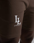 Close-up of LL LYFTLYFE logo printed on right thigh of dark brown shorts.