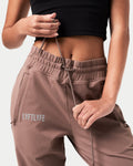 Close-up of the reflective Lyftlyfe logo on the right thigh of women’s brown Breathe Joggers with a thick waistband.
