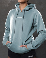 Close-up of center front logo and tonal drawstrings on women’s hoodie in slate color.