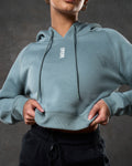 Zoomed-in front view of cropped women’s hoodie in slate color, highlighting logo and stitching.