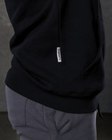 Dominate flag tag detail on black hoodie above kangaroo pocket seam.