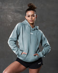 Model in LYFTLYFE women's hoodie in slate and black gym shorts posing against dark wall.