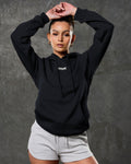 Model in LYFTLYFE women's hoodie in black and gym shorts in light grey posing against dark wall.