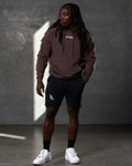 Full-body shot of man in LYFTLYFE brown hoodie and black shorts, standing confidently.
