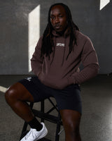 Seated view of man wearing brown LYFTLYFE hoodie and black shorts.