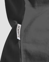 Close-up of the Dominate Adversity flag tag on the dark grey Contour Women’s oversized T-Shirt, stitched on the lower side seam for brand detailing.